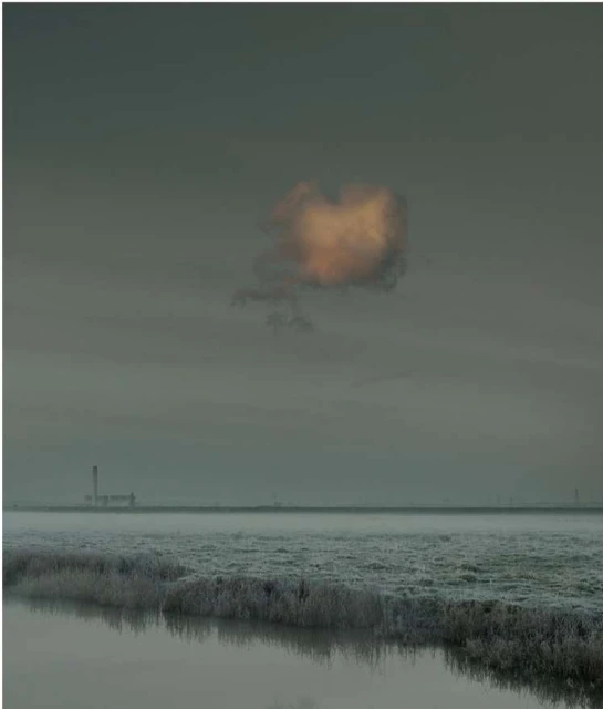 Low-angle landscape captures a cloud illuminated by the sun above a field and calm water, industrial tower in the distant fog.