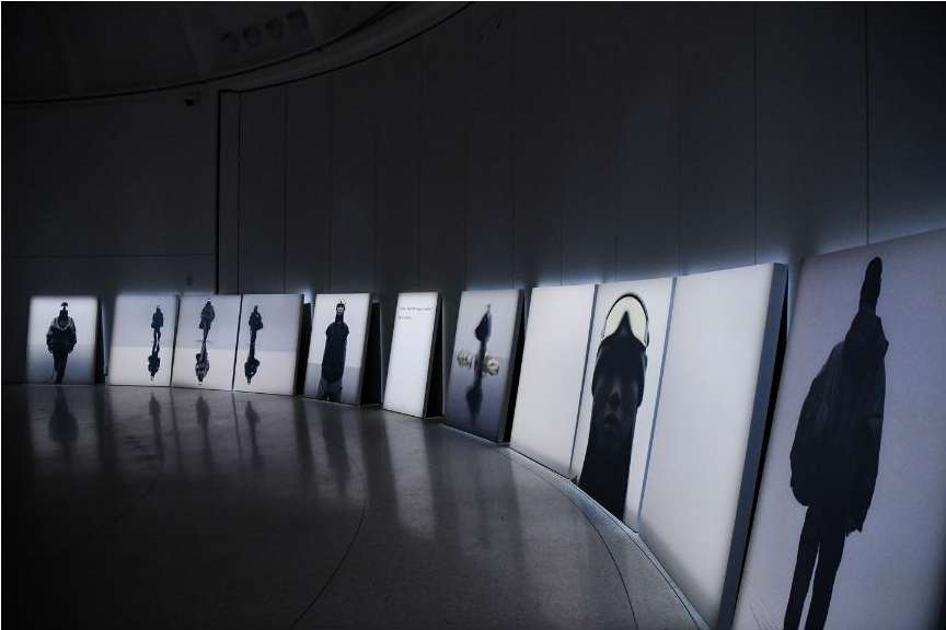 An indoor art installation features a curved row of framed photographs of silhouetted figures in black and white, lit from behind.
