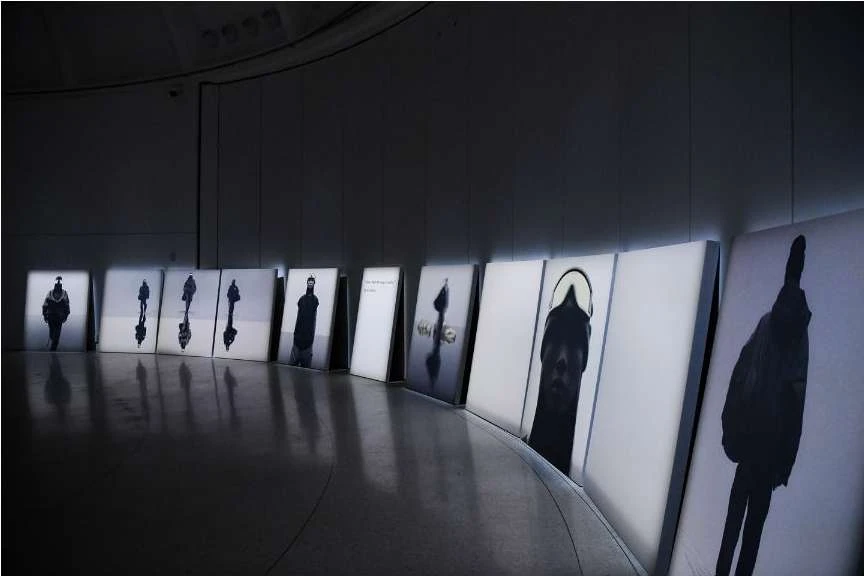 An indoor art installation features a curved row of framed photographs of silhouetted figures in black and white, lit from behind.