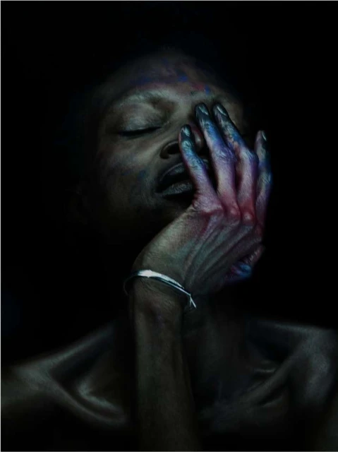 Close-up of an African-descent adult woman with closed eyes, her face partially covered by a hand with a bracelet against a dark backdrop.
