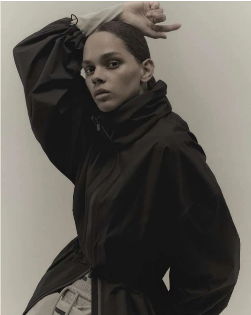 Indoor portrait of a woman in a black jacket with high collar, posing against a light-toned background with hand raised.