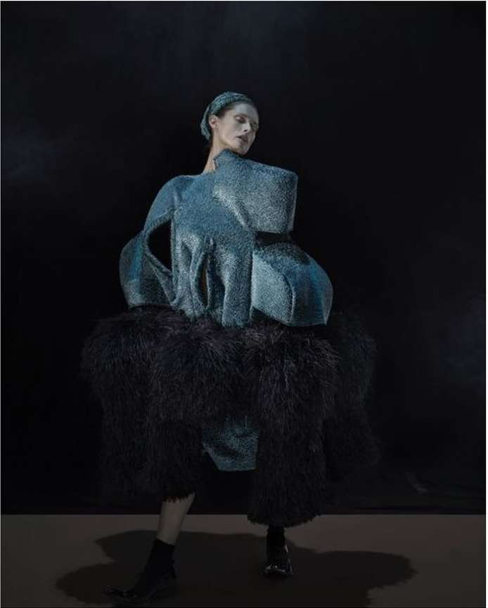 Fashion model poses in an avant-garde blue dress with dark feathered accents against a dark background in a studio setting.