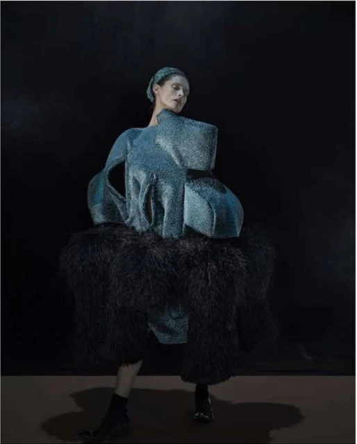Fashion model poses in an avant-garde blue dress with dark feathered accents against a dark background in a studio setting.