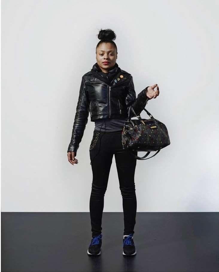Full shot of an African American woman wearing a leather jacket, black pants, and sneakers, holding a satchel-style handbag against a neutral background.