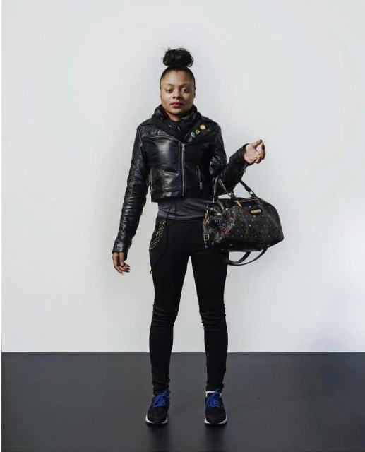Full shot of an African American woman wearing a leather jacket, black pants, and sneakers, holding a satchel-style handbag against a neutral background.