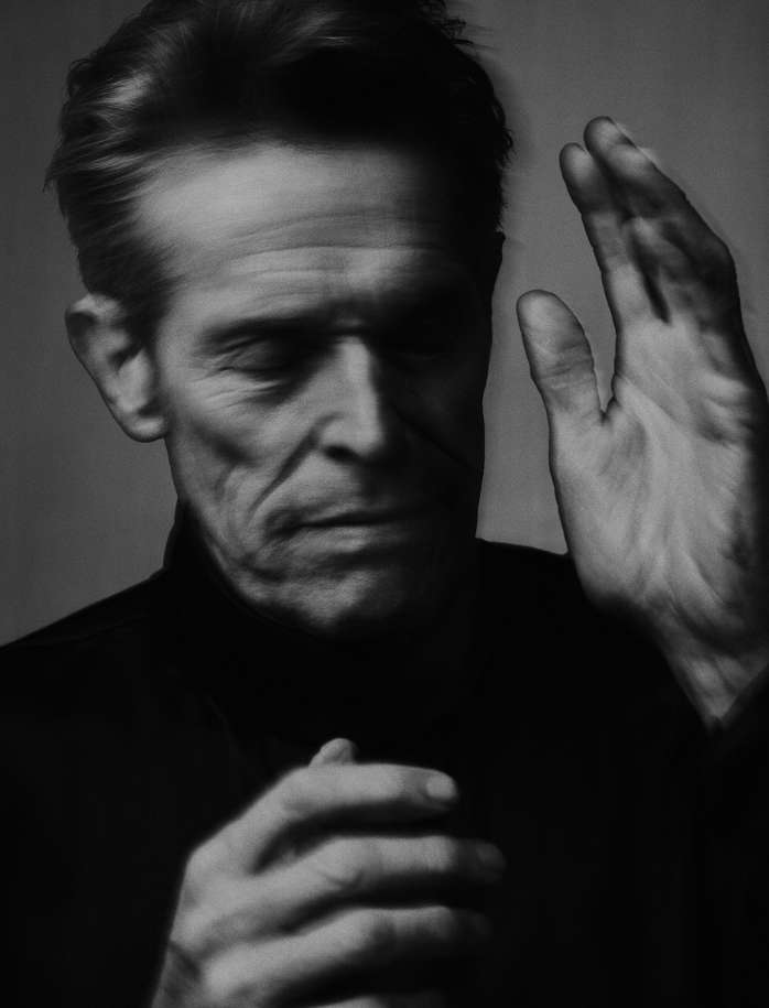 Black and white portrait of a mature man with hands raised, soft lighting, and a neutral background. Facial expression is somber.