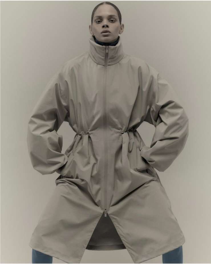 Fashion shot of a model posing in an oversized beige coat with hands in pockets, indoors against a neutral backdrop in a studio setting.