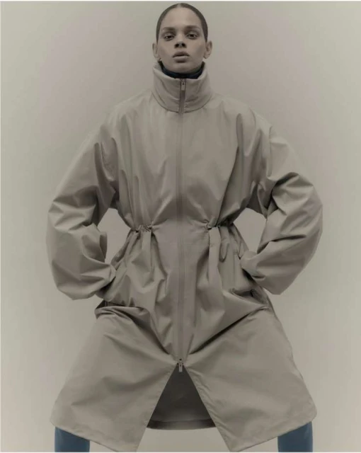 Fashion shot of a model posing in an oversized beige coat with hands in pockets, indoors against a neutral backdrop in a studio setting.