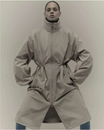Fashion shot of a model posing in an oversized beige coat with hands in pockets, indoors against a neutral backdrop in a studio setting.