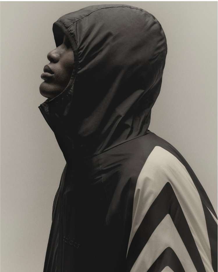Profile of a Black adult male in a hooded windbreaker with chevron design looking up against a plain studio backdrop. Fashion shot.