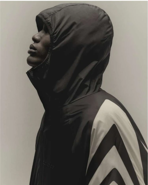 Profile of a Black adult male in a hooded windbreaker with chevron design looking up against a plain studio backdrop. Fashion shot.
