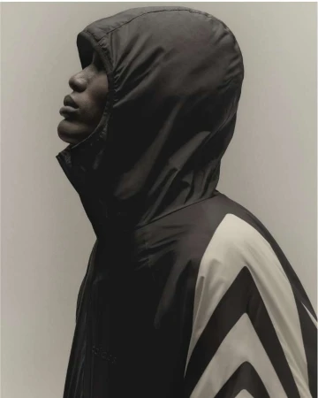 Profile of a Black adult male in a hooded windbreaker with chevron design looking up against a plain studio backdrop. Fashion shot.