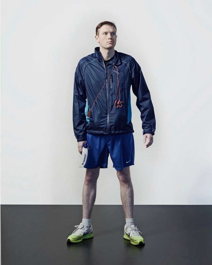 Full shot of a man in athletic wear standing on a black surface against a white background, holding a water bottle.