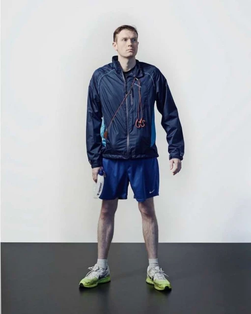 Full shot of a man in athletic wear standing on a black surface against a white background, holding a water bottle.