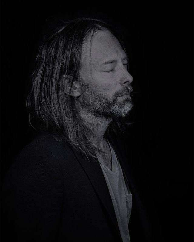 Portrait of Thom Yorke with long hair and beard photographed in low-key lighting against a dark background