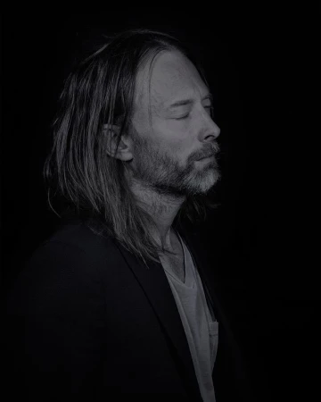 Portrait of Thom Yorke with long hair and beard photographed in low-key lighting against a dark background