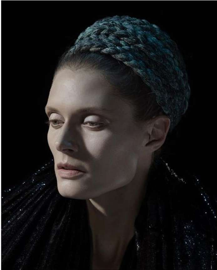 Portrait of a woman wearing a dark blue braided headband and textured dark clothing against a black background.