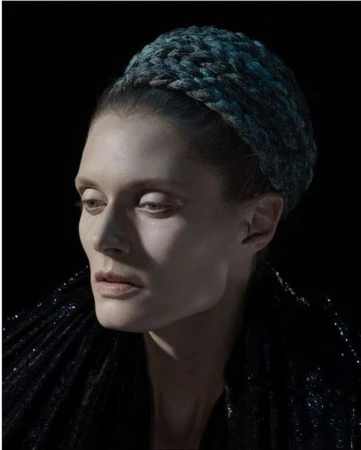 Portrait of a woman wearing a dark blue braided headband and textured dark clothing against a black background.