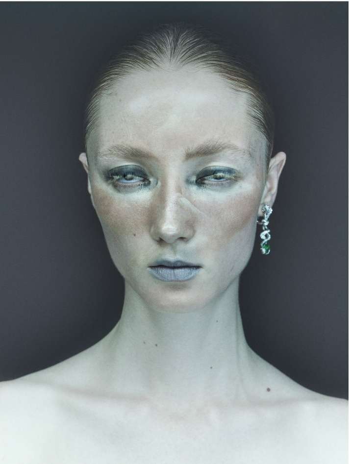 Close-up portrait of a fair-skinned woman with wet slicked-back hair, wearing statement earrings, pale makeup, and looking at the camera.