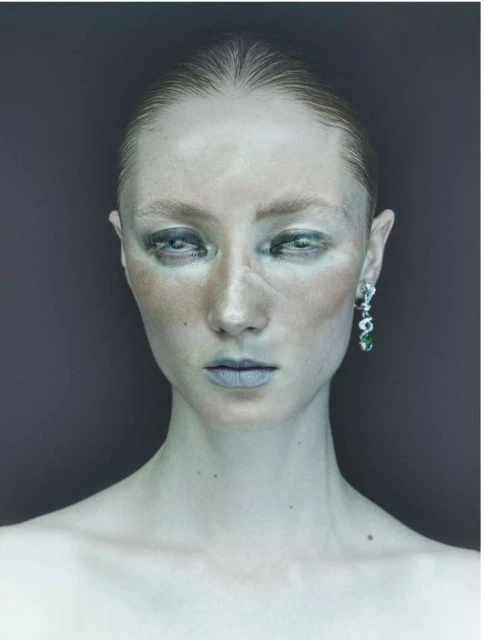 Close-up portrait of a fair-skinned woman with wet slicked-back hair, wearing statement earrings, pale makeup, and looking at the camera.