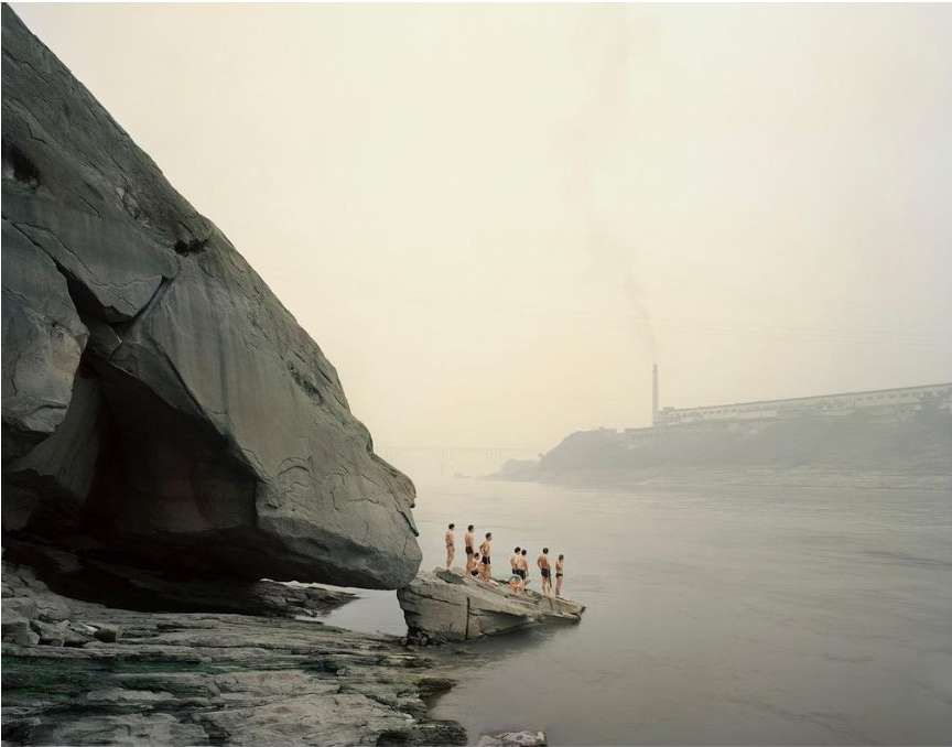 Group of people in swimwear standing on rocks near a large boulder and river, with a factory in the background on a foggy day.
