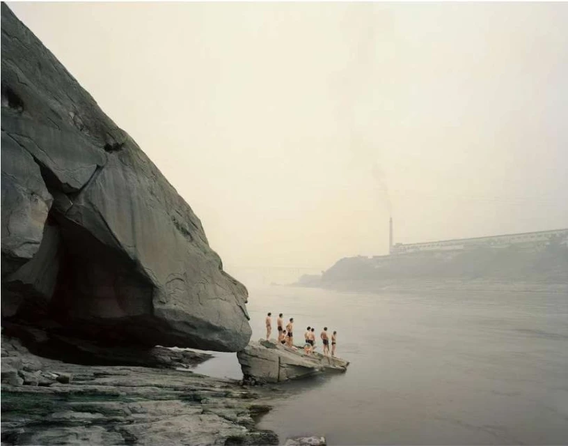 Group of people in swimwear standing on rocks near a large boulder and river, with a factory in the background on a foggy day.