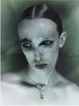 Close-up portrait of a model with slicked-back hair, dramatic dark makeup, and a silver pendant necklace by Nadav Kander.
