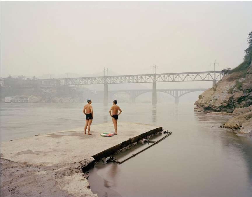 Two shirtless men stand on a concrete platform by a river with bridges in the background under a hazy sky, ready for a swim.