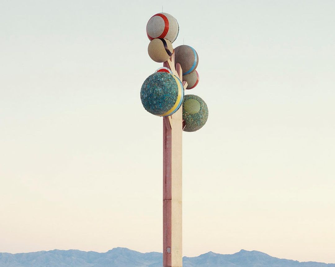 Outdoor art installation featuring stacked colorful spheres atop a tall pole, set against a soft gradient sky and distant mountains.