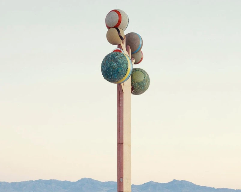 Outdoor art installation featuring stacked colorful spheres atop a tall pole, set against a soft gradient sky and distant mountains.