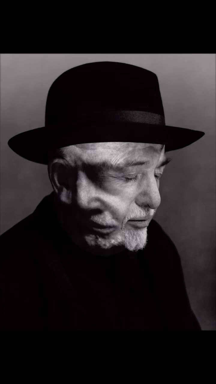 Black and white portrait of an elderly man wearing a black hat, captured in a high-contrast close-up with a soft grey background.