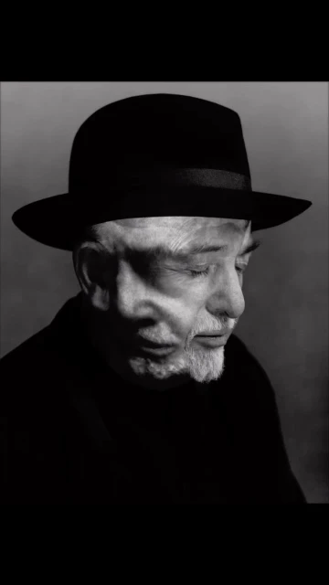 Black and white portrait of an elderly man wearing a black hat, captured in a high-contrast close-up with a soft grey background.