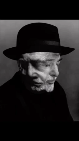 Black and white portrait of an elderly man wearing a black hat, captured in a high-contrast close-up with a soft grey background.