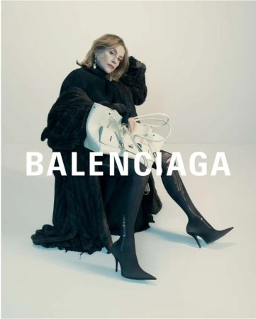 A mature woman seated in a black turtleneck and coat, holding a large white handbag against a plain background