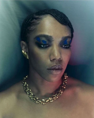 Portrait of a Black woman with braided hair, blue eye makeup, gold chain necklace, and ear cuff against soft background