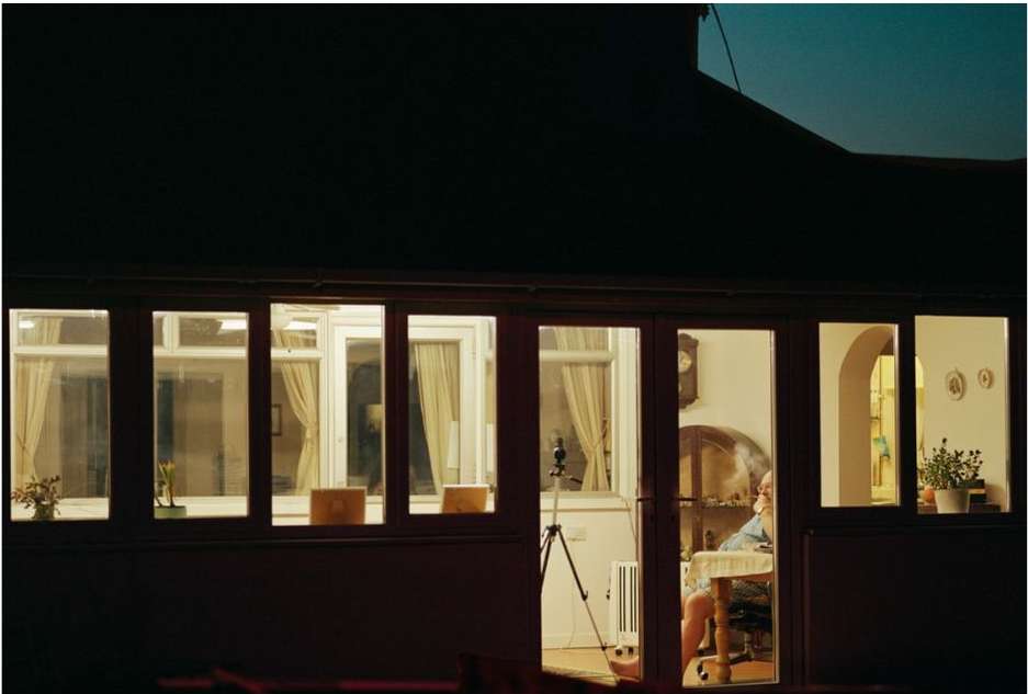 Indoor view through windows showing a person at a table with lamp light, plants, and a tripod visible in a warmly-lit room.