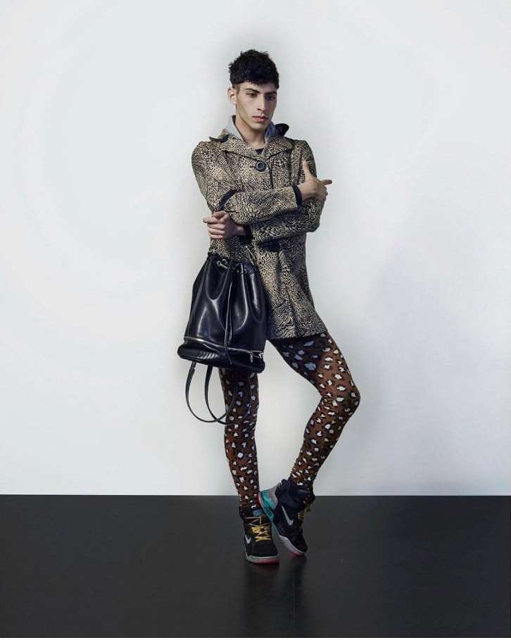 Full-body studio shot of a young adult male wearing a leopard-print coat, patterned tights, sneakers, and carrying a black leather bucket bag.