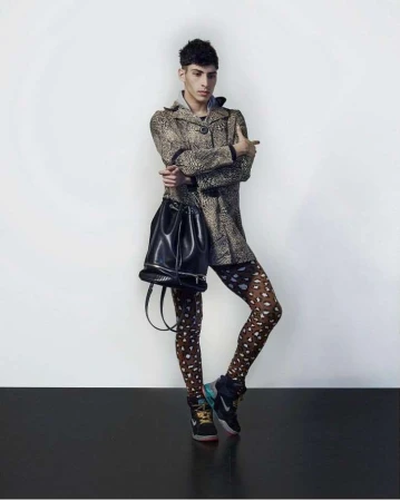 Full-body studio shot of a young adult male wearing a leopard-print coat, patterned tights, sneakers, and carrying a black leather bucket bag.