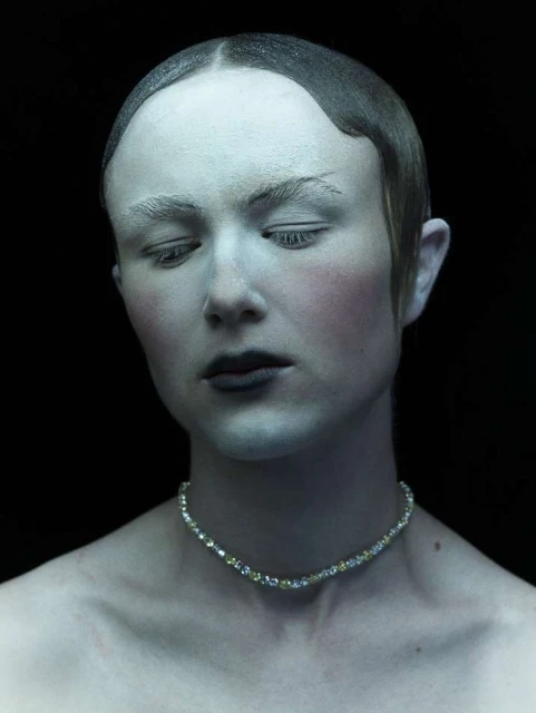 Monochromatic studio portrait of a person with pale skin, dark lips, and a beaded necklace against a solid black backdrop.
