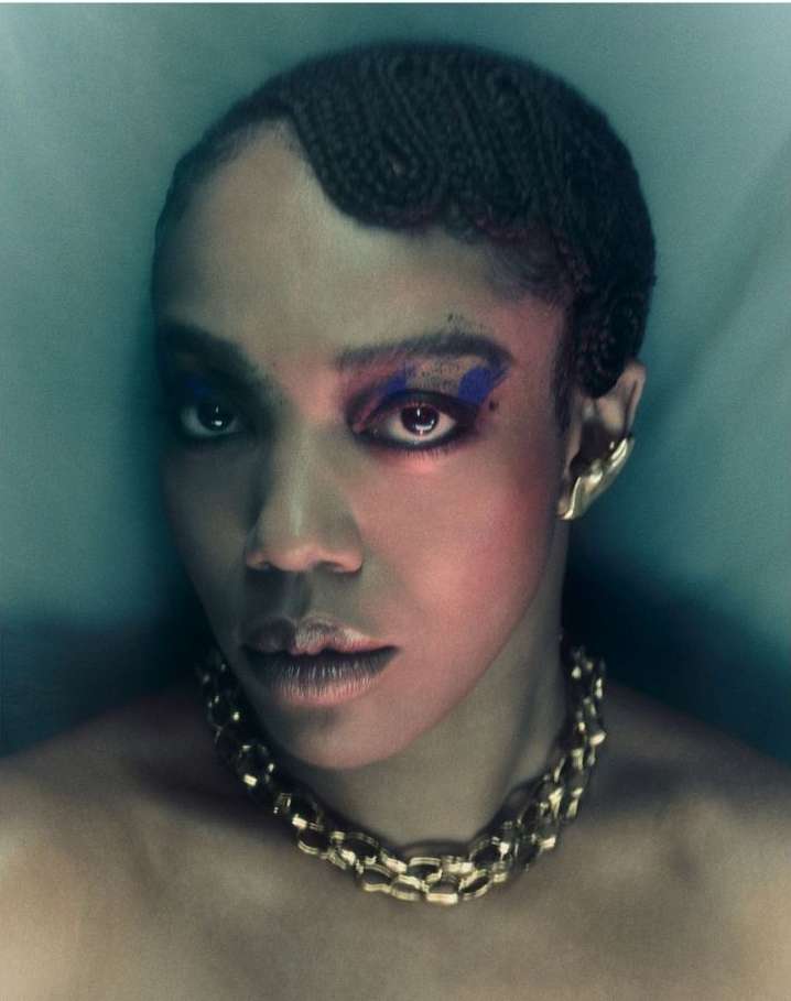 Close-up portrait of a woman with braided hair, dramatic eye makeup, gold jewelry, and direct eye contact against a teal background.