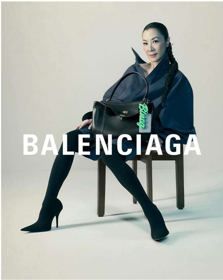 Michelle Yeoh sits on a chair holding a black Balenciaga purse, wearing black leggings and dark blue coat in a fashion studio shot.