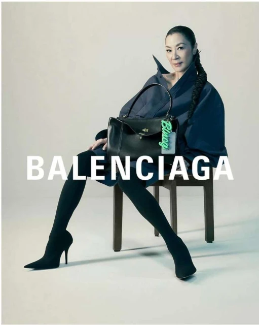 Michelle Yeoh sits on a chair holding a black Balenciaga purse, wearing black leggings and dark blue coat in a fashion studio shot.