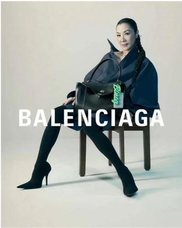 Michelle Yeoh sits on a chair holding a black Balenciaga purse, wearing black leggings and dark blue coat in a fashion studio shot.