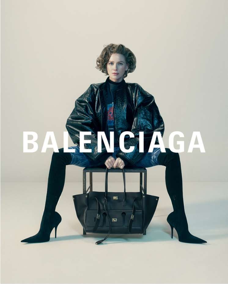 Isabelle Huppert seated on a dark stool holding a black tote bag against a neutral background with Balenciaga typography overlay.