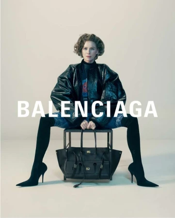 Isabelle Huppert seated on a dark stool holding a black tote bag against a neutral background with Balenciaga typography overlay.