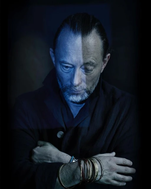 Portrait of a single adult male, wearing a jacket and many bracelets, photographed in a dark indoor environment with blue color grading.