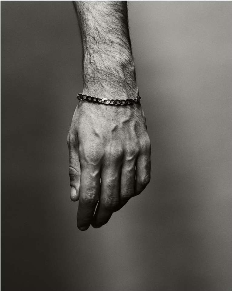 Close-up black and white shot of a male hand with visible veins and hair, wearing a gold chain bracelet, against a neutral background.