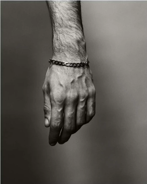 Close-up black and white shot of a male hand with visible veins and hair, wearing a gold chain bracelet, against a neutral background.