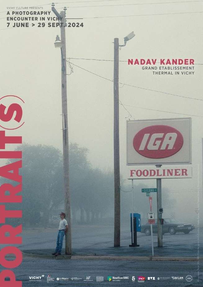 Outdoor advertisement featuring a person leaning against a pole near an IGA grocery store in foggy weather, promoting a photography exhibit.
