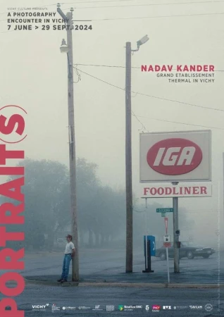 Outdoor advertisement featuring a person leaning against a pole near an IGA grocery store in foggy weather, promoting a photography exhibit.